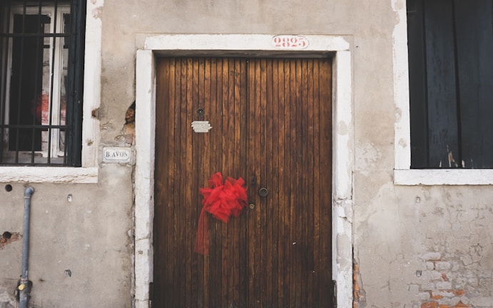 A wooden door with vertical planks is centered between two windows with black metal bars. A vibrant red ribbon is tied to the door handle. The building exterior features an aged, weathered appearance with cracked plaster and exposed bricks. Above the door, a possible address number '9825' is visible, and to the left of the door, there is a street sign labeled 'B AVON'.