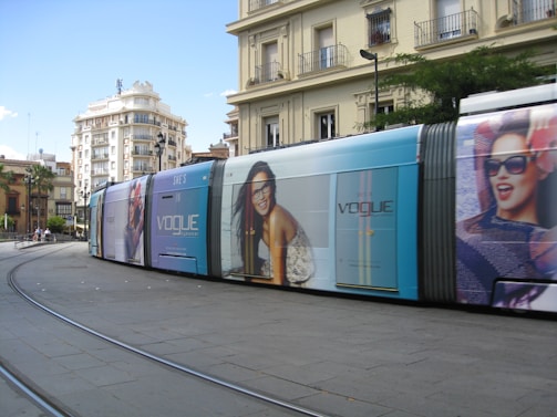 A modern tram with advertisements moves along a curving rail in an urban area. Large posters featuring smiling models with sunglasses are displayed on the side of the tram. The background consists of multi-story buildings with residential balconies and some greenery.