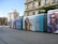 A modern tram with advertisements moves along a curving rail in an urban area. Large posters featuring smiling models with sunglasses are displayed on the side of the tram. The background consists of multi-story buildings with residential balconies and some greenery.