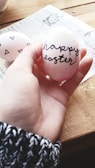 A hand holds an Easter egg with 'happy easter' written in cursive script. The egg is light pink with some darker pink shades and black text. There is another egg with geometric black designs visible in the background, lying on a piece of newspaper on a wooden table. The hand wears a black and white knit sweater.