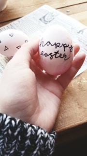 A hand holds an Easter egg with 'happy easter' written in cursive script. The egg is light pink with some darker pink shades and black text. There is another egg with geometric black designs visible in the background, lying on a piece of newspaper on a wooden table. The hand wears a black and white knit sweater.