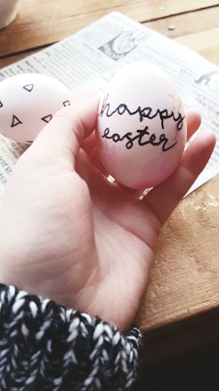 A hand holds an Easter egg with 'happy easter' written in cursive script. The egg is light pink with some darker pink shades and black text. There is another egg with geometric black designs visible in the background, lying on a piece of newspaper on a wooden table. The hand wears a black and white knit sweater.