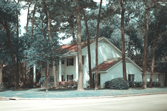 A white two-story house with a red roof, surrounded by tall pine trees. The house is situated in a quiet suburban area, and there is a well-maintained lawn and landscaping with bushes and small trees around it.