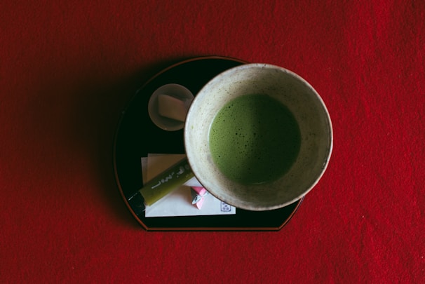 A ceramic bowl containing matcha tea sits on a red surface. The bowl rests on a black tray, accompanied by a small cup with a sugar cube and a wrapped candy. A bamboo stirrer is also placed on the tray.