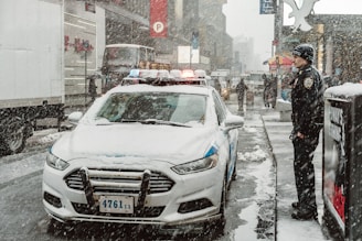 A snowy urban street scene featuring a police car parked on the side of the road. Snow blankets the vehicles and the pavement while a uniformed officer stands nearby. Buildings and pedestrians are visible in the background, creating a bustling city atmosphere despite the weather.