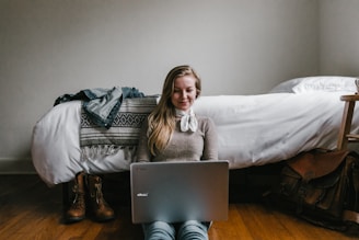 woman sitting beside a bed while using a laptop