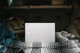 A silver Acer laptop is placed on a patterned blanket in the back of a vehicle. Surrounding it is a denim jacket and a brown backpack, suggesting a casual or travel setting. The interior of the car is visible, indicating it might be parked.