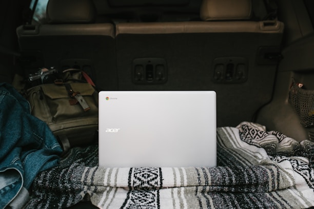 A silver Acer laptop is placed on a patterned blanket in the back of a vehicle. Surrounding it is a denim jacket and a brown backpack, suggesting a casual or travel setting. The interior of the car is visible, indicating it might be parked.
