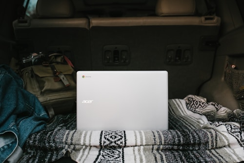 A silver Acer laptop is placed on a patterned blanket in the back of a vehicle. Surrounding it is a denim jacket and a brown backpack, suggesting a casual or travel setting. The interior of the car is visible, indicating it might be parked.