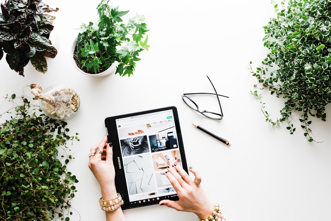 A women with various rings and bracelts working on her tablet around a table of different green plants along with her glasses and pencil.