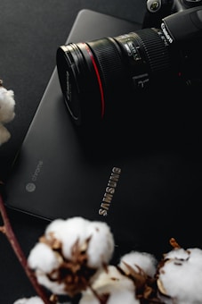 A camera placed on top of a sleek black Samsung Chromebook. Cotton plants are visible in the foreground, adding a contrasting natural element to the tech-themed setup. The camera's branding is prominently displayed, giving it an authoritative presence in the composition.