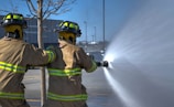 Two firefighters wearing protective gear are using a hose to spray water. The scene takes place outdoors, where a tree and building can be seen in the background. Sunlight glistens off the water spray, creating a dynamic and energetic atmosphere.