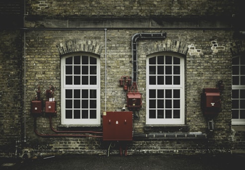 A brick wall facade features two large, arched windows with white frames. Red electrical boxes and piping are mounted on the wall, adding an industrial aspect to the scene. The weathered texture of the bricks provides a historic feel.