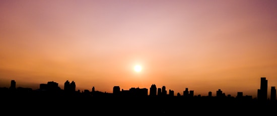 A city skyline silhouetted against a vibrant sunset with hues of orange, pink, and purple. The sun is visible near the center of the image, casting a warm glow over the scene.