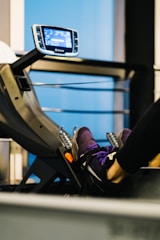 A close-up of a person's feet strapped onto a pedal machine in a gym. The person is wearing purple athletic shoes. A digital display panel is visible in the background, showing various metrics. The environment appears to be an indoor gym with blue and black tones.