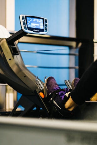 A close-up of a person's feet strapped onto a pedal machine in a gym. The person is wearing purple athletic shoes. A digital display panel is visible in the background, showing various metrics. The environment appears to be an indoor gym with blue and black tones.