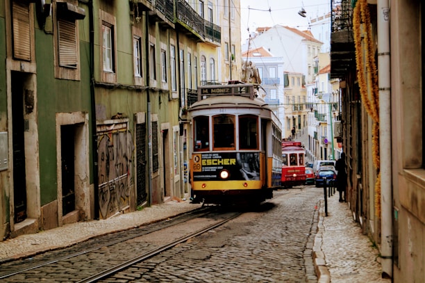 A narrow street in a European city is lined with old buildings, showcasing distinct architectural styles. A vintage tram approaches, carrying passengers as it navigates the tram tracks embedded in the cobblestone road. The street is busy with activity, as people walk along the sidewalks and other vehicles follow the tram.