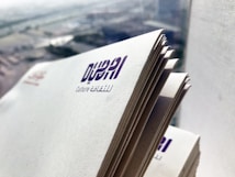 A stack of papers with the logo and text 'Dubai Culture' printed on each page. The image is taken close-up with an urban cityscape visible in the blurred background.