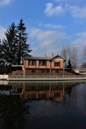 A brick house with a stone foundation is situated by a calm body of water, reflecting its image. The sky is partly cloudy with a vibrant blue hue. Tall trees, both evergreen and leafless, surround the property, creating a serene setting.