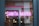 Neon pink lights spell out the words 'Santa Maria' in an interior setting, with reflections on a glass surface displaying elements of the outside street and inside lighting fixtures. Warm hanging light bulbs and a background with tiles add to the ambience.