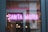 Neon pink lights spell out the words 'Santa Maria' in an interior setting, with reflections on a glass surface displaying elements of the outside street and inside lighting fixtures. Warm hanging light bulbs and a background with tiles add to the ambience.