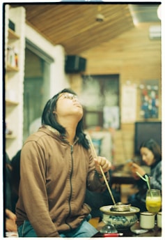 A person wearing glasses and a brown hoodie is sitting indoors at a table, holding a long stick and leaning back slightly with their eyes closed, exhaling smoke upwards. On the table, there is a floral-patterned pot with a small fire, a plant, a glass of yellowish drink with a straw, and several other objects. In the background, another person is focused on their phone, sitting against a wooden wall with various items and decorations visible.