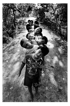 A group of children is standing in a line on a dirt path, each leaning their head to one side. They appear joyful and are all looking directly at the camera. The path is surrounded by trees and bushes, creating a natural, serene environment with dappled sunlight creating patterns on the ground.