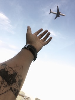 A hand reaches towards the sky with a tattoo of a world map on the forearm. An airplane flies overhead, set against a backdrop of clear blue sky.