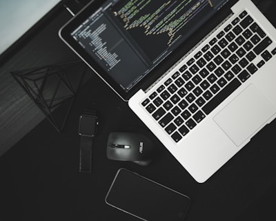 A workspace setup with a laptop displaying code on its screen, accompanied by a wireless mouse, a smartwatch, and a smartphone on a dark surface. A geometric wireframe object is placed nearby, adding a modern touch.
