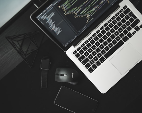 A workspace setup with a laptop displaying code on its screen, accompanied by a wireless mouse, a smartwatch, and a smartphone on a dark surface. A geometric wireframe object is placed nearby, adding a modern touch.