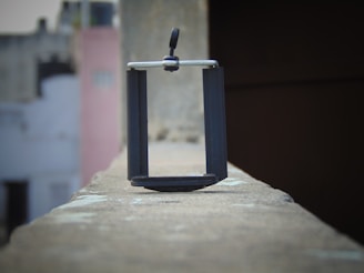 A black phone holder is placed on the edge of a rooftop wall. The background is blurry, displaying buildings with faded colors, including white, gray, and pale pink. The image is captured at an angle that highlights the focus on the phone holder, giving a sense of depth and perspective.
