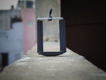 A black phone holder is placed on the edge of a rooftop wall. The background is blurry, displaying buildings with faded colors, including white, gray, and pale pink. The image is captured at an angle that highlights the focus on the phone holder, giving a sense of depth and perspective.