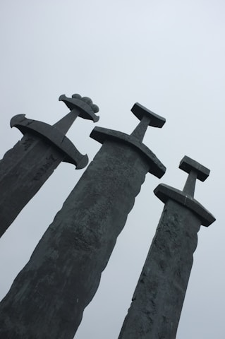 Three large swords stand upright, embedded in the ground with their blades pointing towards the sky. The swords have distinct, oversized hilts and are made of a weathered, dark metal. The background is a pale, overcast sky, adding a cool, subdued atmosphere to the scene.