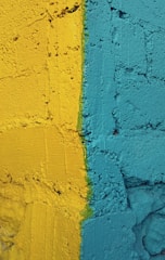 A textured wall featuring two bold colors, with a distinct dividing line separating a vibrant yellow from a striking teal. The wall's surface has a rough, uneven texture, adding depth to the color contrast.