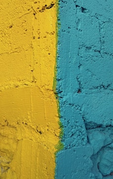 A textured wall featuring two bold colors, with a distinct dividing line separating a vibrant yellow from a striking teal. The wall's surface has a rough, uneven texture, adding depth to the color contrast.