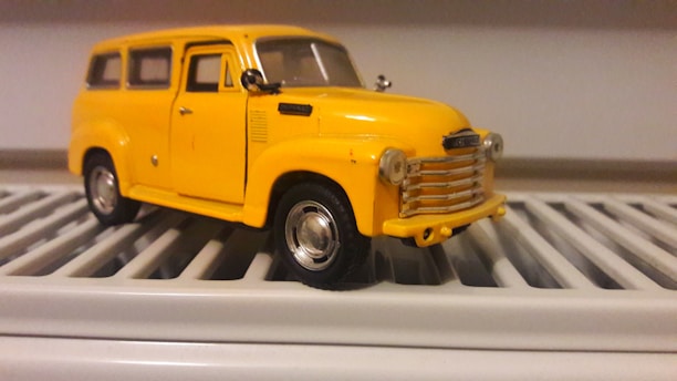 A detailed miniature model of a vintage yellow car is placed on a white, slatted surface. The car features intricate details, including side mirrors, a chrome grille, and realistic wheels.