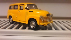 A detailed miniature model of a vintage yellow car is placed on a white, slatted surface. The car features intricate details, including side mirrors, a chrome grille, and realistic wheels.
