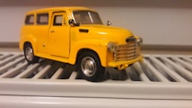 A detailed miniature model of a vintage yellow car is placed on a white, slatted surface. The car features intricate details, including side mirrors, a chrome grille, and realistic wheels.