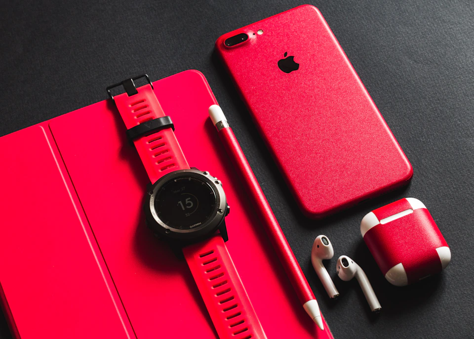The image presents a collection of everyday objects, including an Apple iPhone with a red case and a white earbud, a black watch with a red strap, a red pen, a red pencil, and a red eraser. The objects are arranged on a black surface against a gray background, creating a visually striking contrast between the vibrant colors of the items and their surroundings.