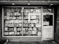 A black and white exterior view of an old bookstore featuring large display windows filled with an assortment of books. Signs on the window advertise ‘Old Books’ and ‘Literary History Walking Tour’. A variety of book titles are visible, and the shop door on the right displays more signage including a notice for a literary walking tour.
