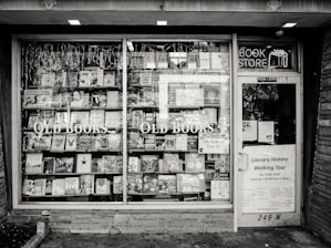 A black and white exterior view of an old bookstore featuring large display windows filled with an assortment of books. Signs on the window advertise ‘Old Books’ and ‘Literary History Walking Tour’. A variety of book titles are visible, and the shop door on the right displays more signage including a notice for a literary walking tour.