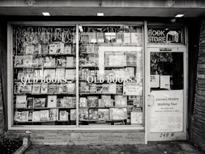 A black and white exterior view of an old bookstore featuring large display windows filled with an assortment of books. Signs on the window advertise ‘Old Books’ and ‘Literary History Walking Tour’. A variety of book titles are visible, and the shop door on the right displays more signage including a notice for a literary walking tour.