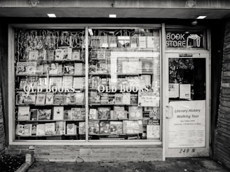 A black and white exterior view of an old bookstore featuring large display windows filled with an assortment of books. Signs on the window advertise ‘Old Books’ and ‘Literary History Walking Tour’. A variety of book titles are visible, and the shop door on the right displays more signage including a notice for a literary walking tour.