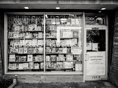 A black and white exterior view of an old bookstore featuring large display windows filled with an assortment of books. Signs on the window advertise ‘Old Books’ and ‘Literary History Walking Tour’. A variety of book titles are visible, and the shop door on the right displays more signage including a notice for a literary walking tour.