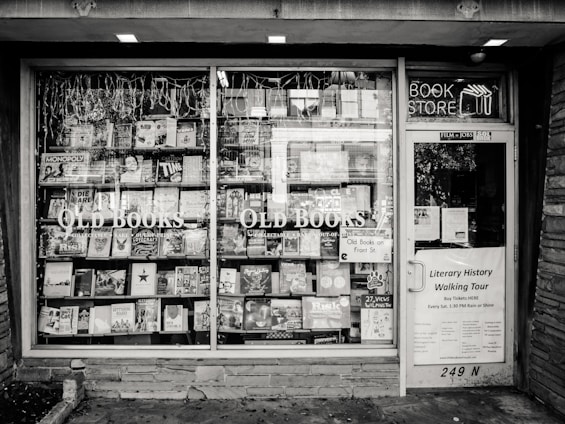 A black and white exterior view of an old bookstore featuring large display windows filled with an assortment of books. Signs on the window advertise ‘Old Books’ and ‘Literary History Walking Tour’. A variety of book titles are visible, and the shop door on the right displays more signage including a notice for a literary walking tour.