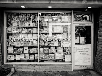 A black and white exterior view of an old bookstore featuring large display windows filled with an assortment of books. Signs on the window advertise ‘Old Books’ and ‘Literary History Walking Tour’. A variety of book titles are visible, and the shop door on the right displays more signage including a notice for a literary walking tour.