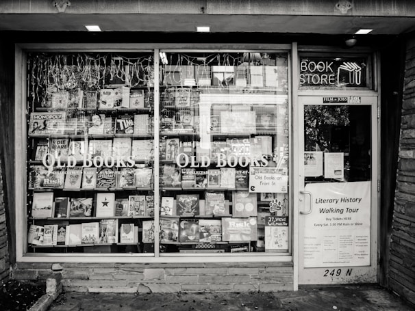 A black and white exterior view of an old bookstore featuring large display windows filled with an assortment of books. Signs on the window advertise ‘Old Books’ and ‘Literary History Walking Tour’. A variety of book titles are visible, and the shop door on the right displays more signage including a notice for a literary walking tour.