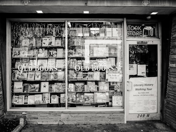 A black and white exterior view of an old bookstore featuring large display windows filled with an assortment of books. Signs on the window advertise &lsquo;Old Books&rsquo; and &lsquo;Literary History Walking Tour&rsquo;. A variety of book titles are visible, and the shop door on the right displays more signage including a notice for a literary walking tour.