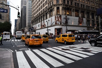 Yellow taxis are driving through a busy city intersection surrounded by tall buildings. A large department store is on the corner with a billboard ad above it. Pedestrians are crossing the street, and a traffic light is visible.