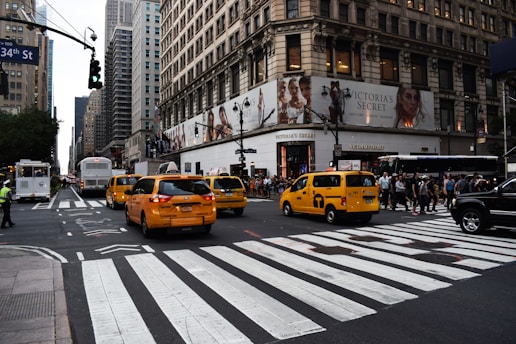Yellow taxis are driving through a busy city intersection surrounded by tall buildings. A large department store is on the corner with a billboard ad above it. Pedestrians are crossing the street, and a traffic light is visible.